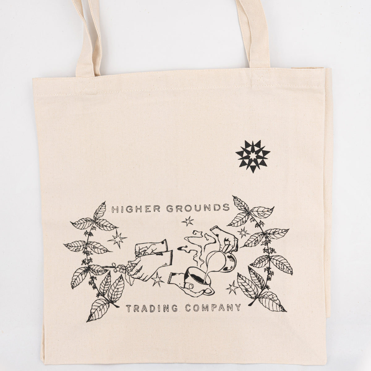 A natural canvas tote bag with black printed graphics and text reading 'HIGHER GROUNDS TRADING COMPANY' on a white background with illustrated image of hands, coffee plants, and a latte.