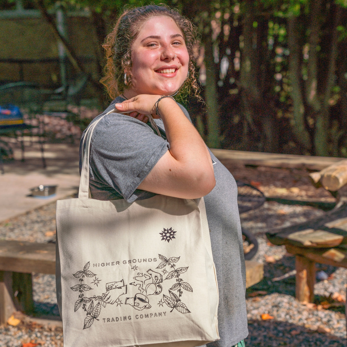 Person holding a natural canvas tote bag with black printed graphics and text reading 'HIGHER GROUNDS TRADING COMPANY' on a white background with illustrated image of hands, coffee plants, and a latte.